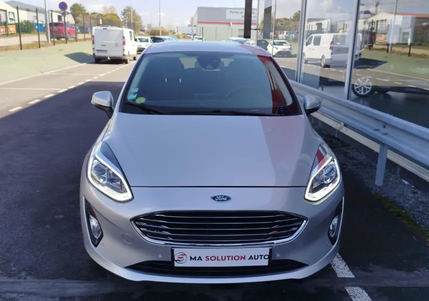 Vue avant d'une Ford Fiesta gris lunaire 2019 avec phares LED allumés, garée devant un bâtiment vitré.