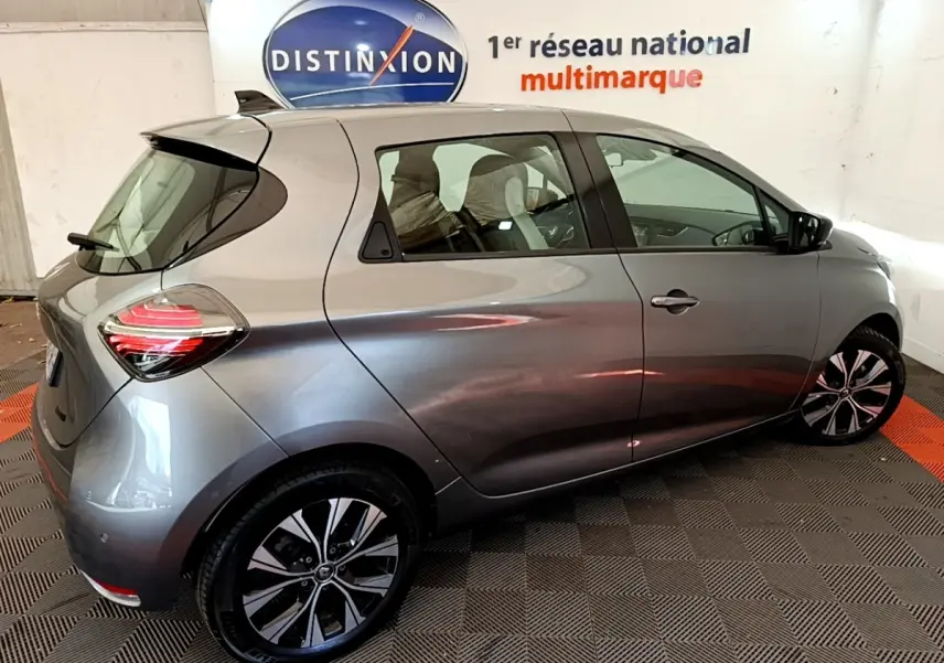 Vue 3/4 arrière droite d'une Renault ZOE gris clair électrique, avec jantes noires et logo Renault visible sur la roue.