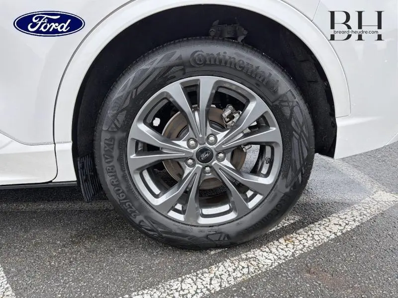 Gros plan sur la roue avant droite du Ford Kuga blanc Premium Platinum, avec jante alu et logo Ford central visible.
