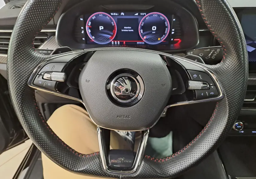 Vue rapprochée du volant perforé noir avec surpiqûres rouges et logo Skoda, tableau de bord digital en arrière-plan.