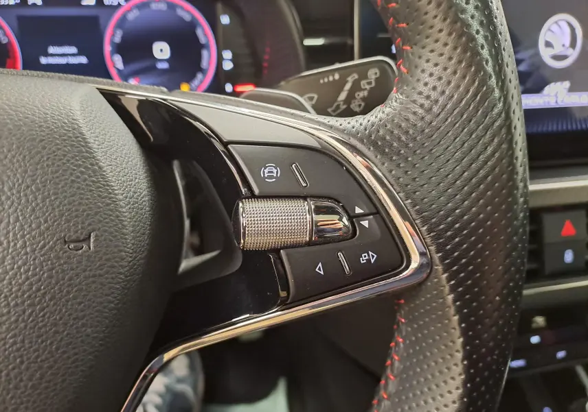 Gros plan sur la partie droite du volant perforé avec surpiqûres rouges du Skoda Scala noir Magic nacré, tableau de bord numérique en arrière-plan.