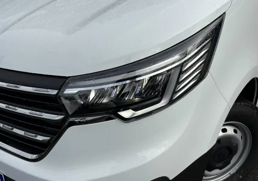 Gros plan sur le phare avant droit et la calandre chromée du Renault Trafic Fourgon blanc glacier, avec gouttes de pluie visibles.