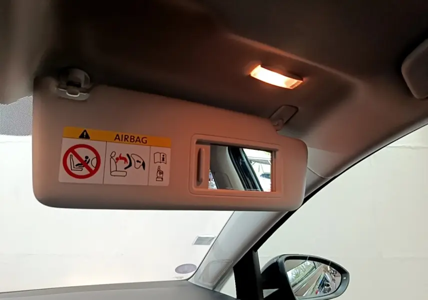 Vue intérieure côté conducteur sur le pare-soleil beige avec miroir et avertissement airbag, rétroviseur noir visible.