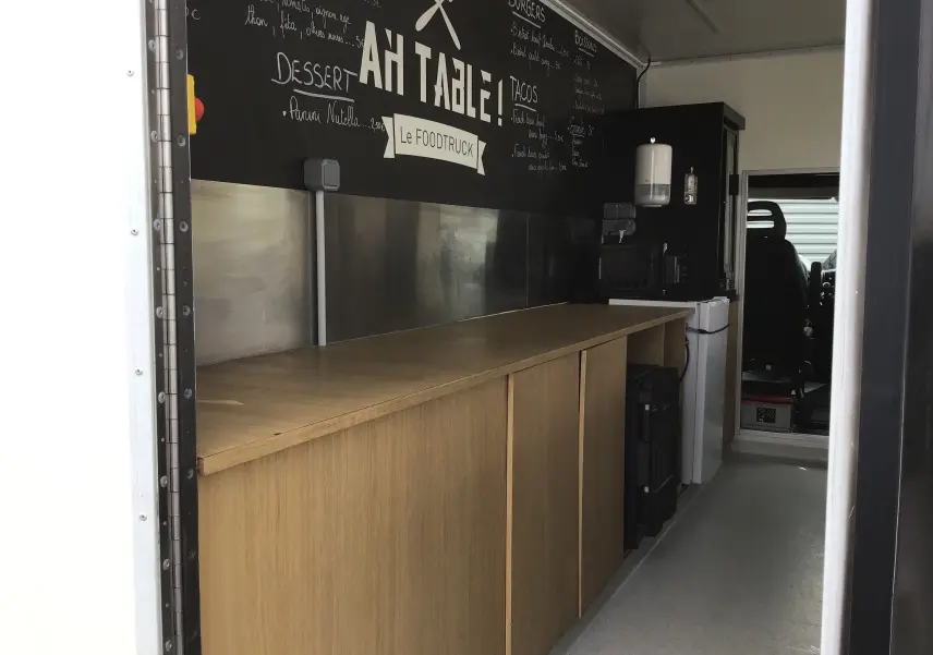 Intérieur du food truck Citroën Jumper gris, vue latérale droite sur plan de travail en bois et mur noir avec menu écrit.