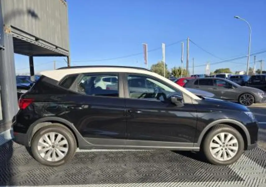 Profil côté gauche d'une SEAT Arona 2024 noir minuit avec toit blanc contrasté, stationnée en extérieur sous un ciel bleu.