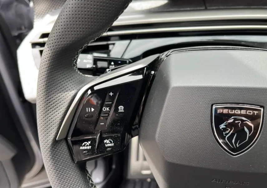 Gros plan sur le volant cuir perforé de la Peugeot 5008 Hybrid GT, avec commandes multifonctions et logo lion central.