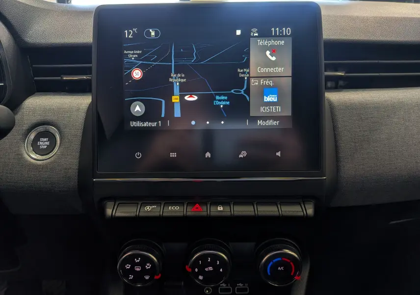 Vue intérieure centrée sur l’écran tactile du tableau de bord de la Renault Clio V gris Rafale avec commandes clim et bouton start visible.