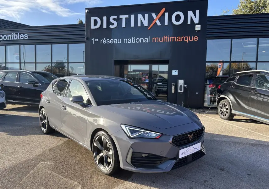 Vue 3/4 avant d'une CUPRA Leon gris Graphene avec jantes alliage noires devant un showroom Distinxion.