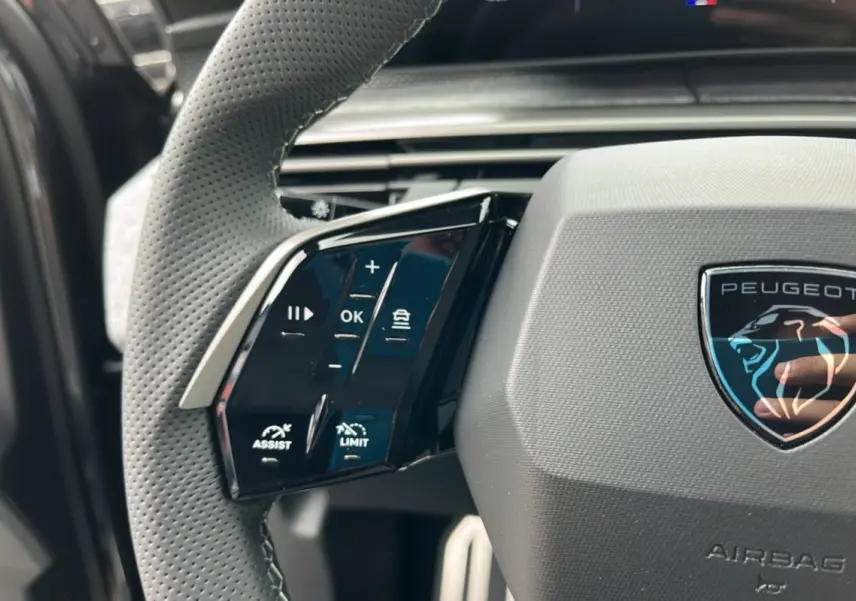 Gros plan sur le volant cuir perforé côté gauche du Peugeot 5008 Hybrid gris titane avec logo Peugeot central.