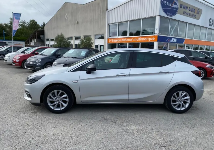 Profil côté gauche d'une Opel Astra Sovereign Silver 2021 garée devant un garage multimarque avec jantes alliage 16 pouces.