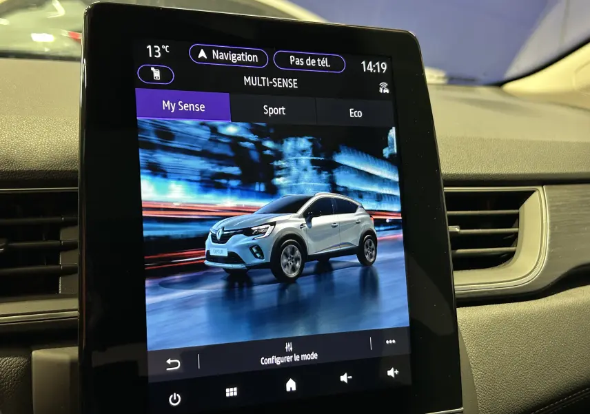 Écran tactile intérieur montrant un Renault Captur blanc nacré en 3/4 avant sur fond urbain flou.