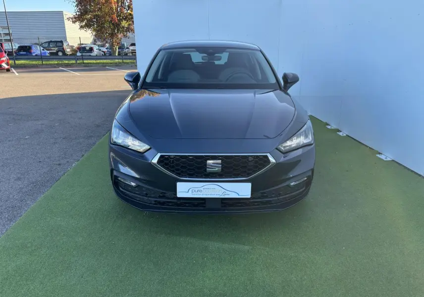 Vue frontale d'une SEAT Leon IV gris magnetic garée sur un sol vert avec calandre hexagonale et phares LED allumés.