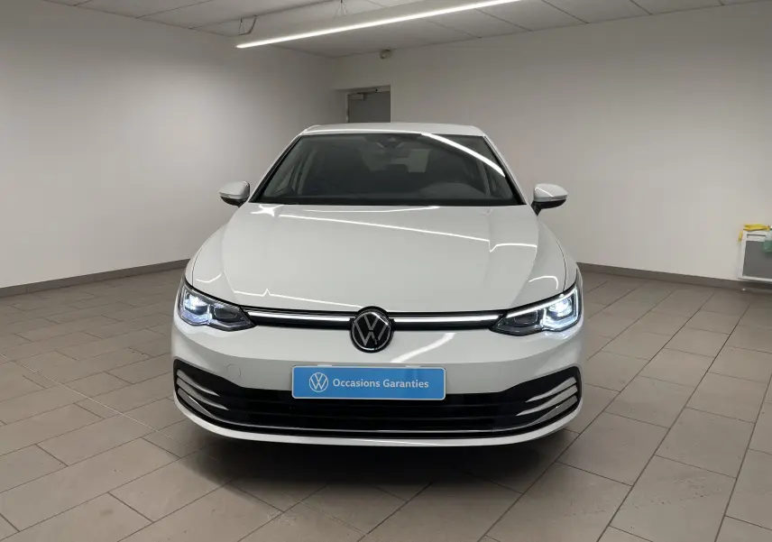 Volkswagen Golf 1.5 eTSI blanc pur vue de face en intérieur avec phares allumés et logo VW central.