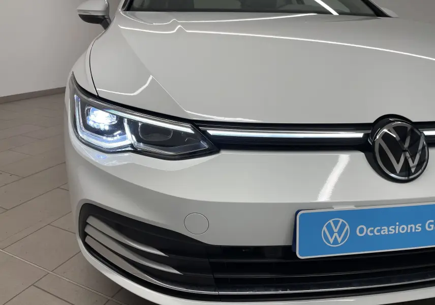 Gros plan sur l'avant gauche d'une Volkswagen Golf 1.5 eTSI blanche avec phare LED allumé et calandre noire.