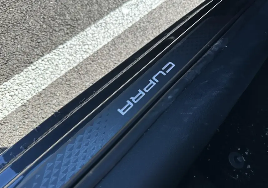 Gros plan sur le seuil de porte côté gauche avec logo CUPRA sur fond noir texturé, intérieur sombre visible.
