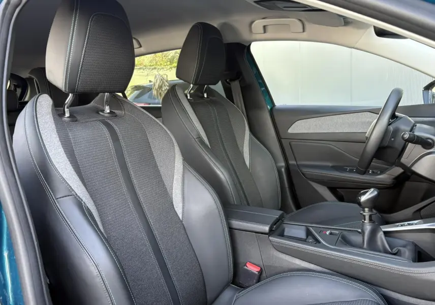 Vue intérieure côté conducteur de la Peugeot 308 2024, sièges tri matières noirs avec surpiqûres bleues et levier de vitesses manuel.