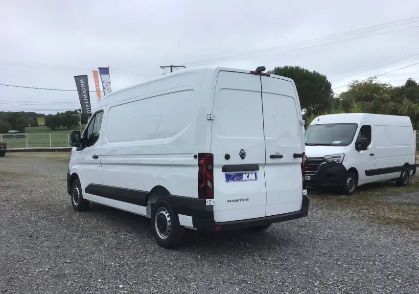 Fourgon Renault Master blanc vu en 3/4 arrière droit sur un parking extérieur avec un autre utilitaire en arrière-plan.
