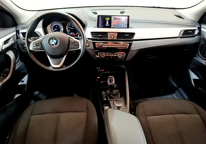 Vue intérieure frontale du tableau de bord et volant cuir noir de la BMW X2 sDrive18i 2023 avec écran central affichant l'aide au stationnement.