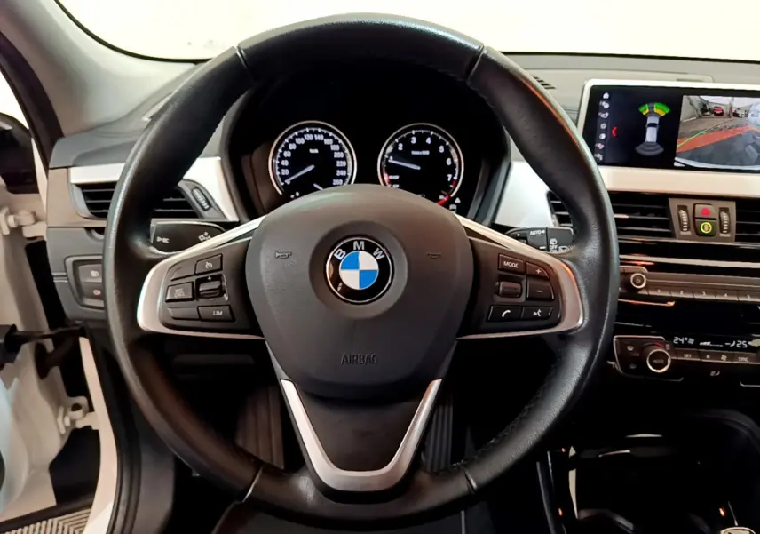Vue intérieure centrée sur le volant cuir de la BMW X2 blanc, avec tableau de bord et écran d'aide au stationnement visibles.