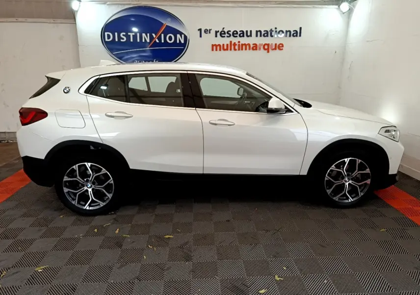 BMW X2 blanc vue de profil côté gauche dans un showroom avec jantes alliage et logo sur l'aile arrière.