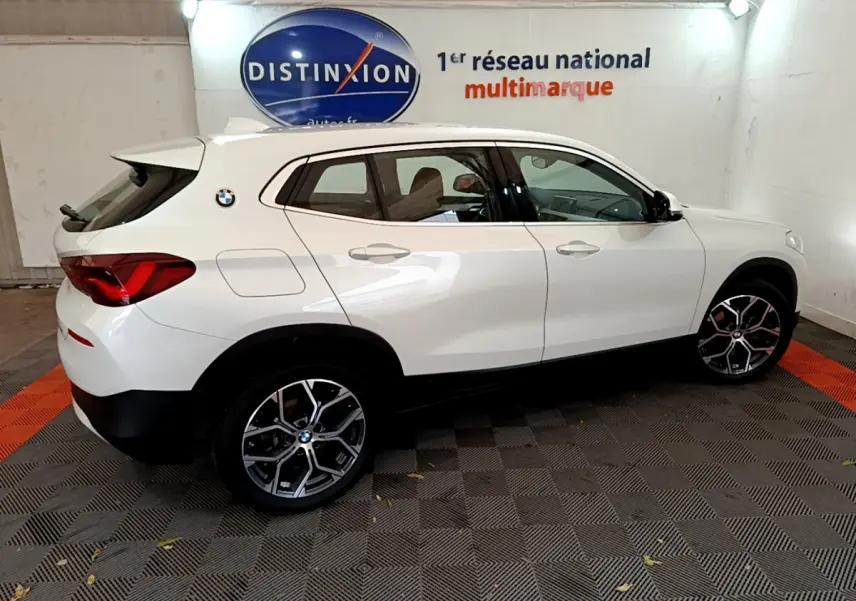 BMW X2 blanc vu de profil côté gauche dans un showroom avec jantes alliage et logo distinctif sur l'aile arrière.