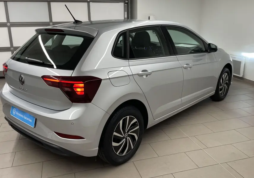 Vue 3/4 arrière droite d'une Volkswagen Polo 1.0 TSI 95 Life 2023 en peinture métallisée reflet d'argent avec feux arrière allumés.