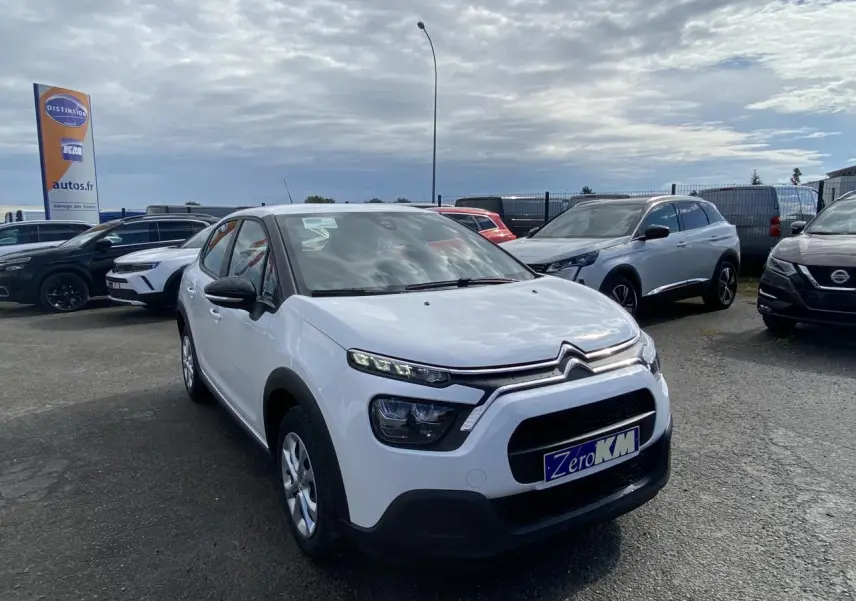 Citroën C3 blanc vue 3/4 avant droit, avec calandre noire et enjoliveurs 15 pouces sur parking extérieur.