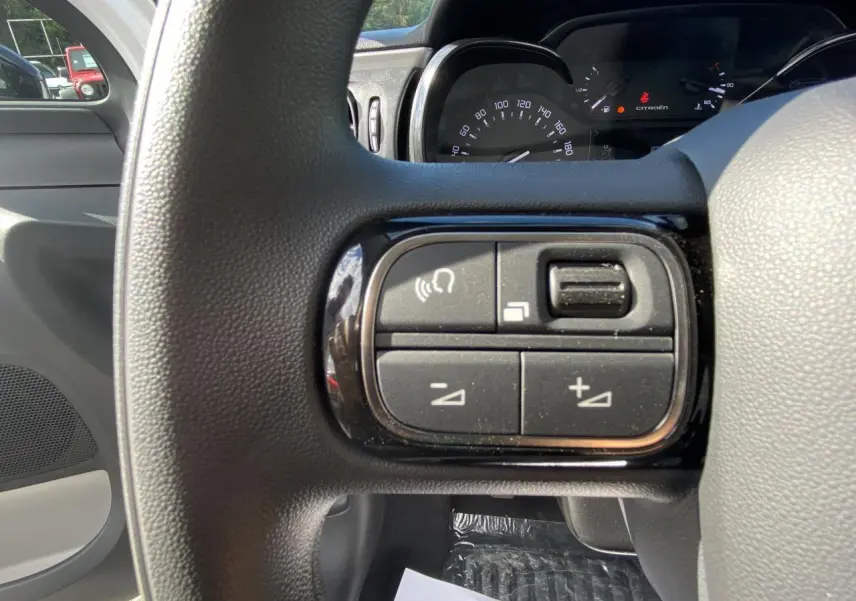 Gros plan sur les commandes audio au volant noir d'une Citroën C3 blanche, tableau de bord et compteur visibles en arrière-plan.