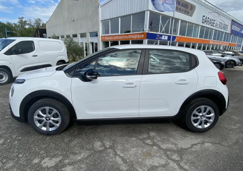 Profil côté gauche d'une Citroën C3 blanche 2022 avec enjoliveurs 15 pouces et rétroviseurs noirs devant un garage.