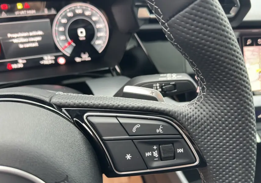 Gros plan sur le volant cuir perforé de l'Audi A3 2025 avec commandes audio et palettes au volant visibles.