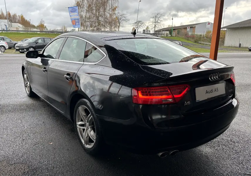 Audi A5 Sportback noire vue en 3/4 arrière droit sous la pluie, avec feux arrière allumés et jantes alliage.