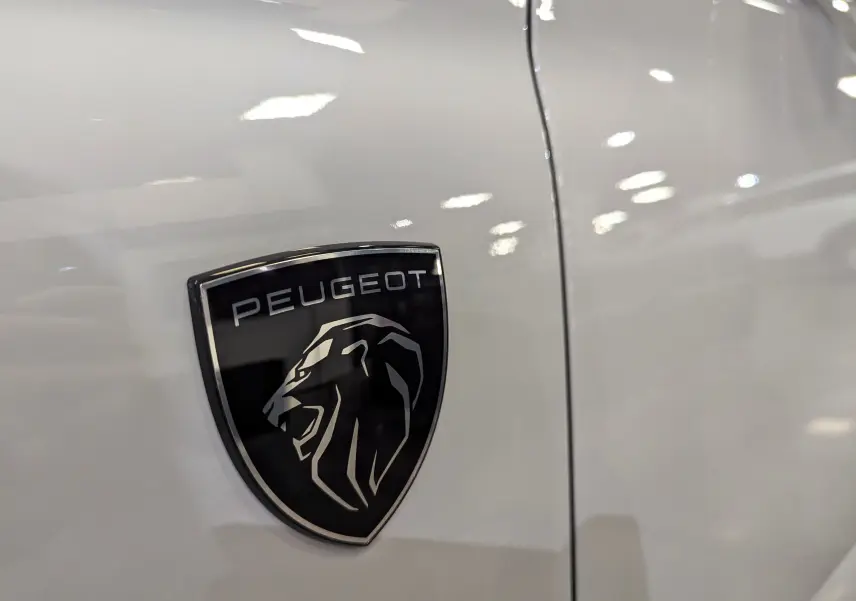 Gros plan sur l'écusson noir et argenté Peugeot fixé sur la carrosserie blanche Okénite d'une 308 2024.