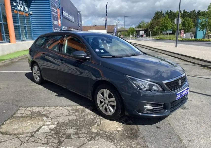 Peugeot 308 SW gris hurricane vue 3/4 avant droit, jantes alliage 16 pouces, stationnée devant un bâtiment commercial.