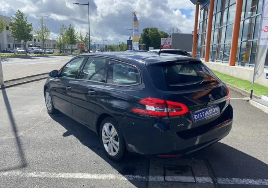 Vue 3/4 arrière droite d'une Peugeot 308 SW gris hurricane garée devant un bâtiment vitré par temps nuageux.