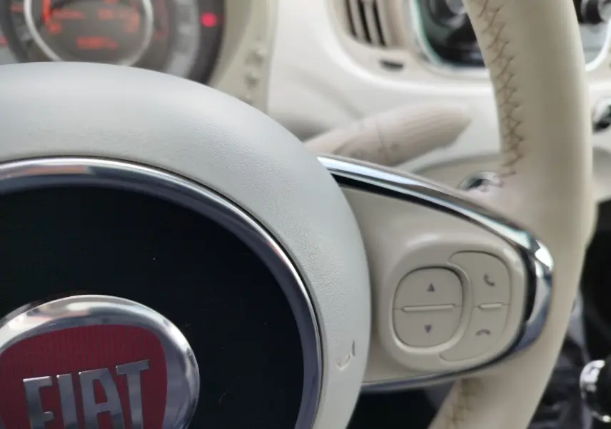 Gros plan sur le volant beige clair de la Fiat 500 Lounge 2017 avec commandes intégrées et logo FIAT rouge au centre.
