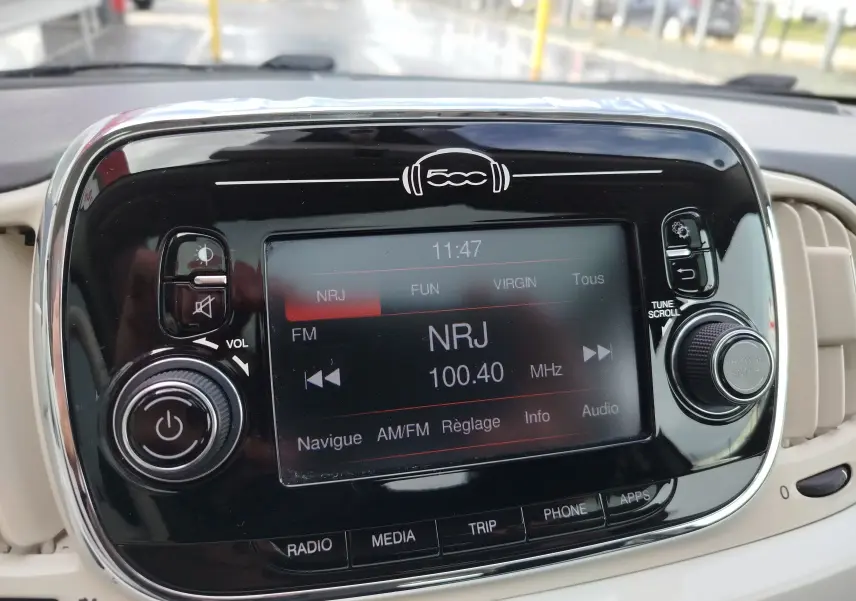 Vue rapprochée de l’écran multimédia noir brillant de la Fiat 500 blanche, affichant la radio NRJ à 100.40 MHz.