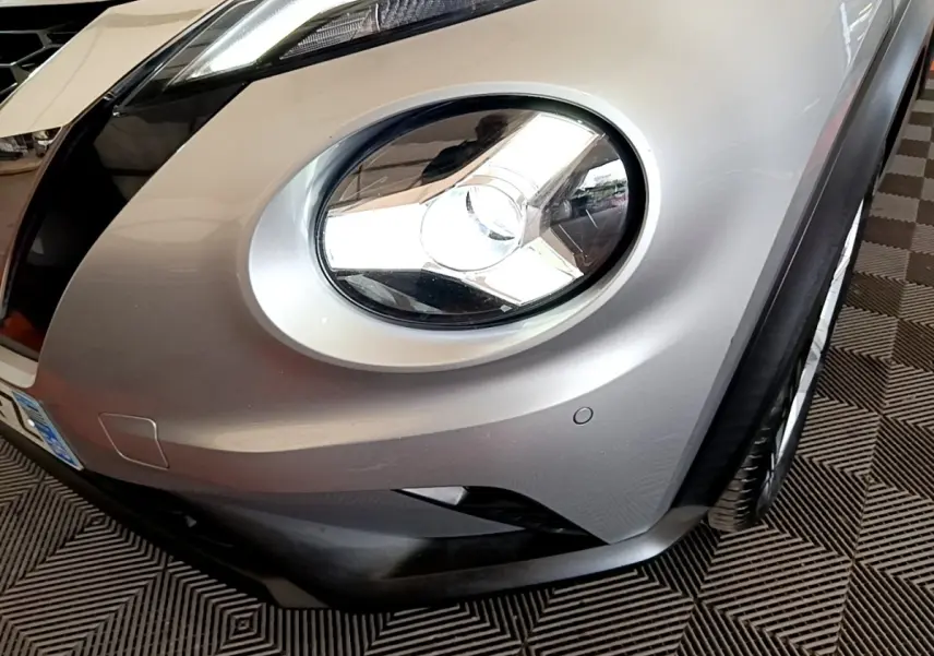 Gros plan sur le phare avant droit du Nissan Juke gris clair, mettant en valeur le design arrondi et le pare-chocs.
