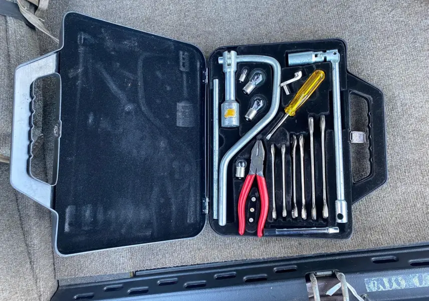 Boîte à outils ouverte avec pinces rouges et tournevis jaune, posée sur la moquette marron d'une Jaguar XJ6 Série 3 1985.