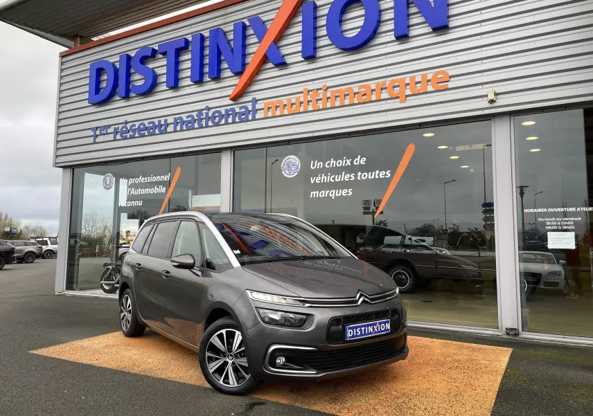 Citroën Grand C4 Picasso gris en 3/4 avant droit, stationné devant un showroom Distinxion avec reflets sur la carrosserie.