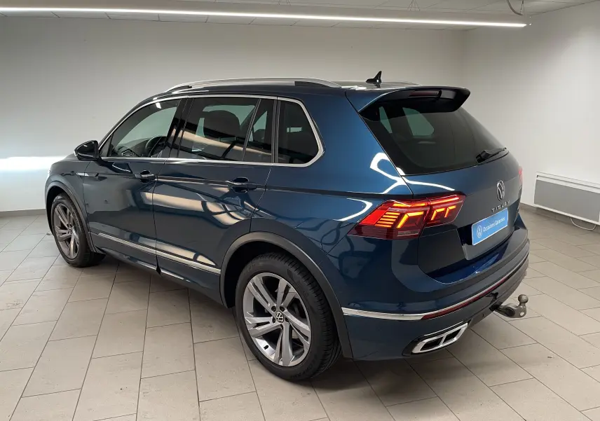 Vue 3/4 arrière droite du Volkswagen Tiguan 2024 en bleu Nightshade avec attelage escamotable et feux allumés.