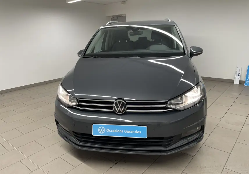 Vue de face d'un Volkswagen Touran gris dauphin 2024 avec phares allumés dans un showroom intérieur.