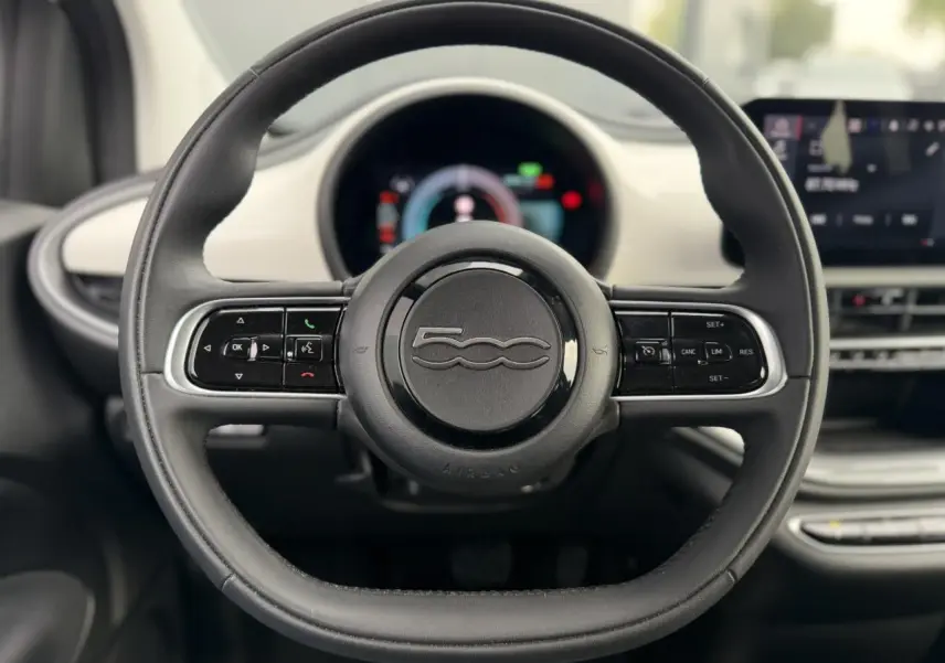 Vue rapprochée du volant noir de la Fiat 500 électrique 2024, avec commandes intégrées et tableau de bord numérique flou.