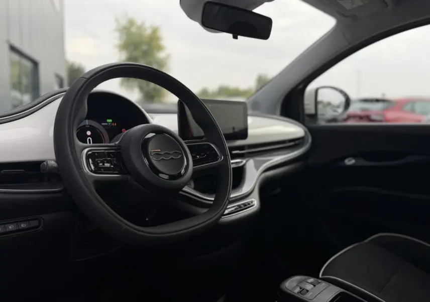 Vue intérieure côté conducteur de la Fiat 500 électrique blanche 2024, avec volant, tableau de bord numérique et écran central.