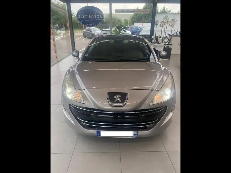 Vue avant d'une Peugeot RCZ gris métallisé 2010 avec phares allumés dans un showroom lumineux.