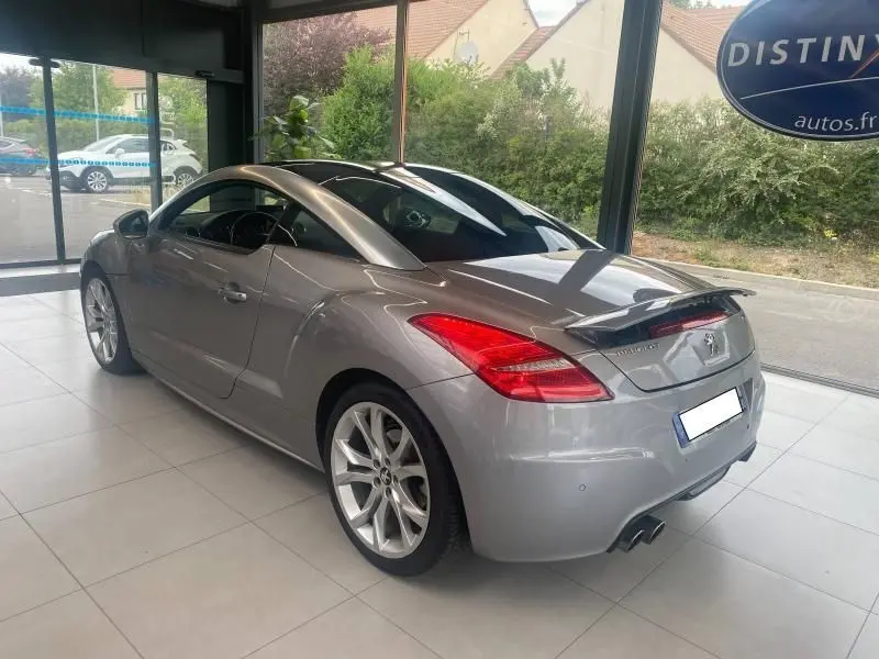 Peugeot RCZ gris vue 3/4 arrière droit en showroom, avec jantes alliage et aileron déployé visible.