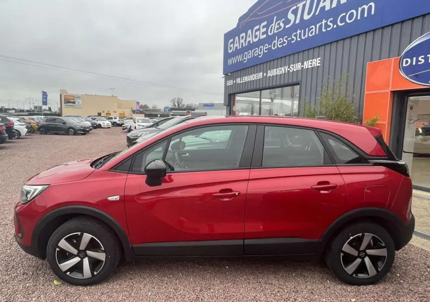 Profil côté gauche d'un Opel Crossland rouge métallisé 2022 garé devant un garage avec jantes noires et argentées.