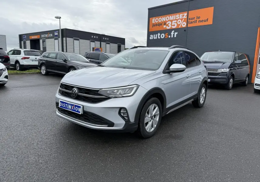 Volkswagen Taigo gris reflet d'argent métallisé en 3/4 avant droit sur parking devant un bâtiment commercial.