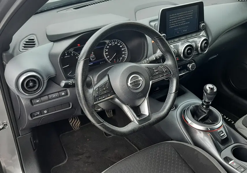 Vue intérieure côté conducteur du Nissan Juke gris 2020, volant cuir, tableau de bord avec écran tactile et levier de vitesse manuel.