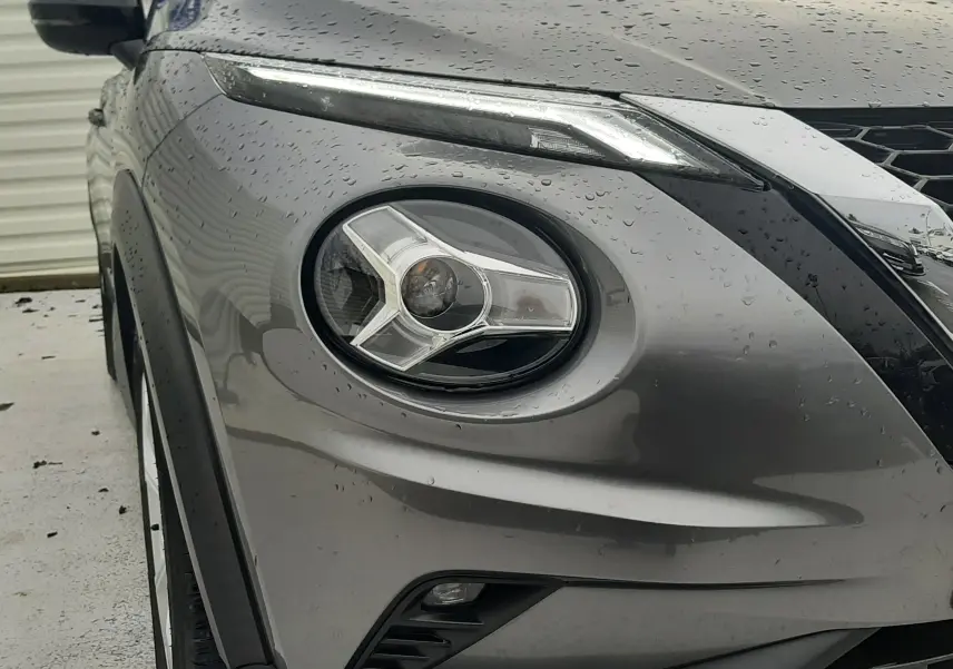 Gros plan sur l'avant gauche gris d'un Nissan Juke 2020, mettant en valeur son phare rond et sa calandre noire mouillée.