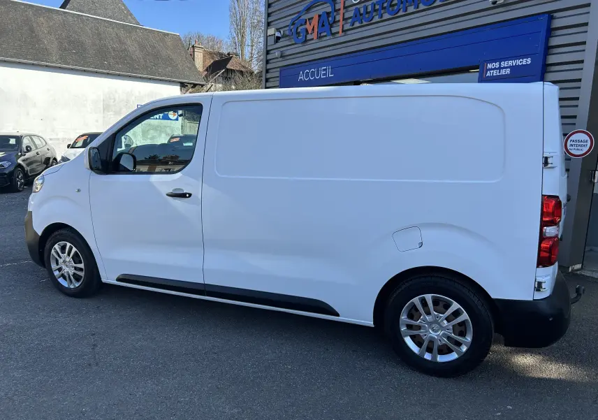 Vue de profil côté gauche d’un fourgon Citroën Jumpy blanc avec attelage, stationné devant un garage automobile.